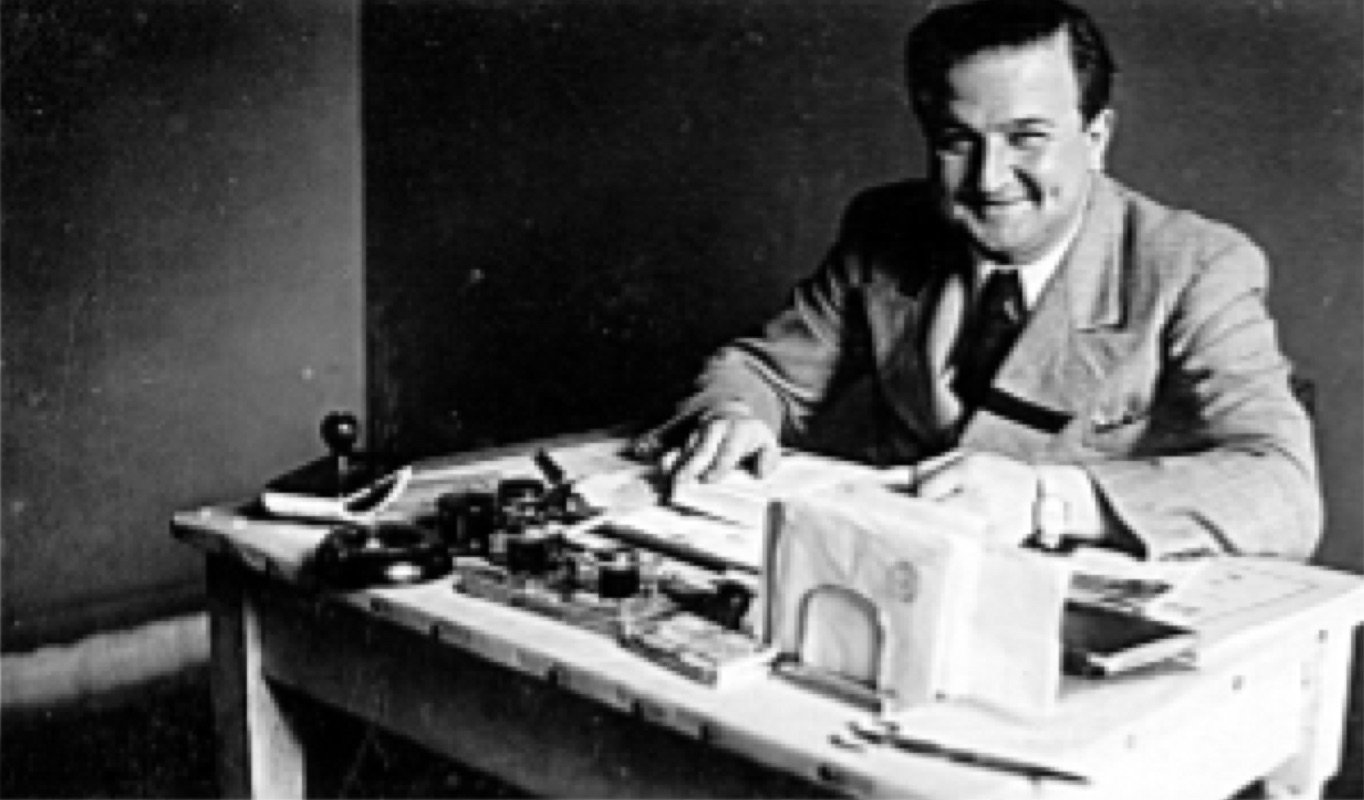 Louis Sader, sorridente, sentado à sua mesa de trabalho em foto antiga em preto e branco