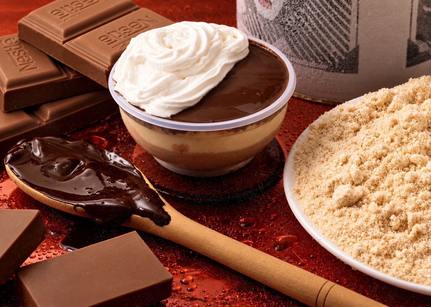 Pote de sorvete com calda de chocolate e chantilly ao lado de barras de chocolate, de uma colher de pau e de um prato cheio de farofa doce